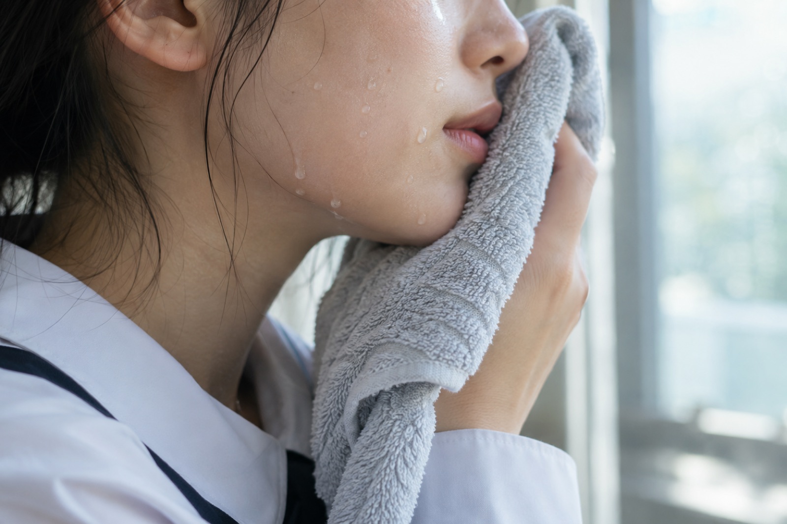 窓辺で白い制服のシャツを着た学生が、水滴のついた頬と口元にグレーのタオルを当てている横顔