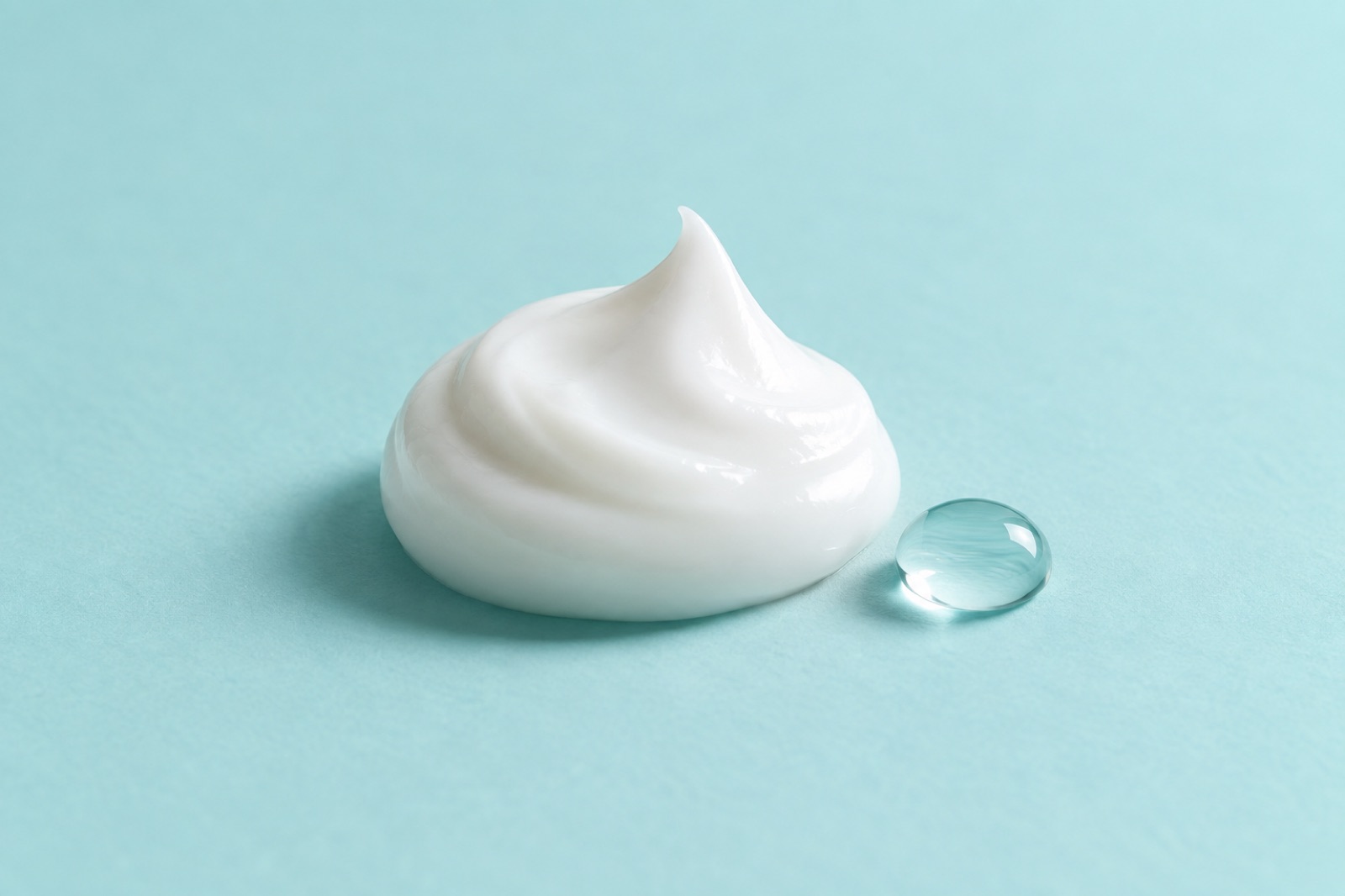 A single peaked dollop of white cream on a sky-blue surface with one small clear water drop beside it