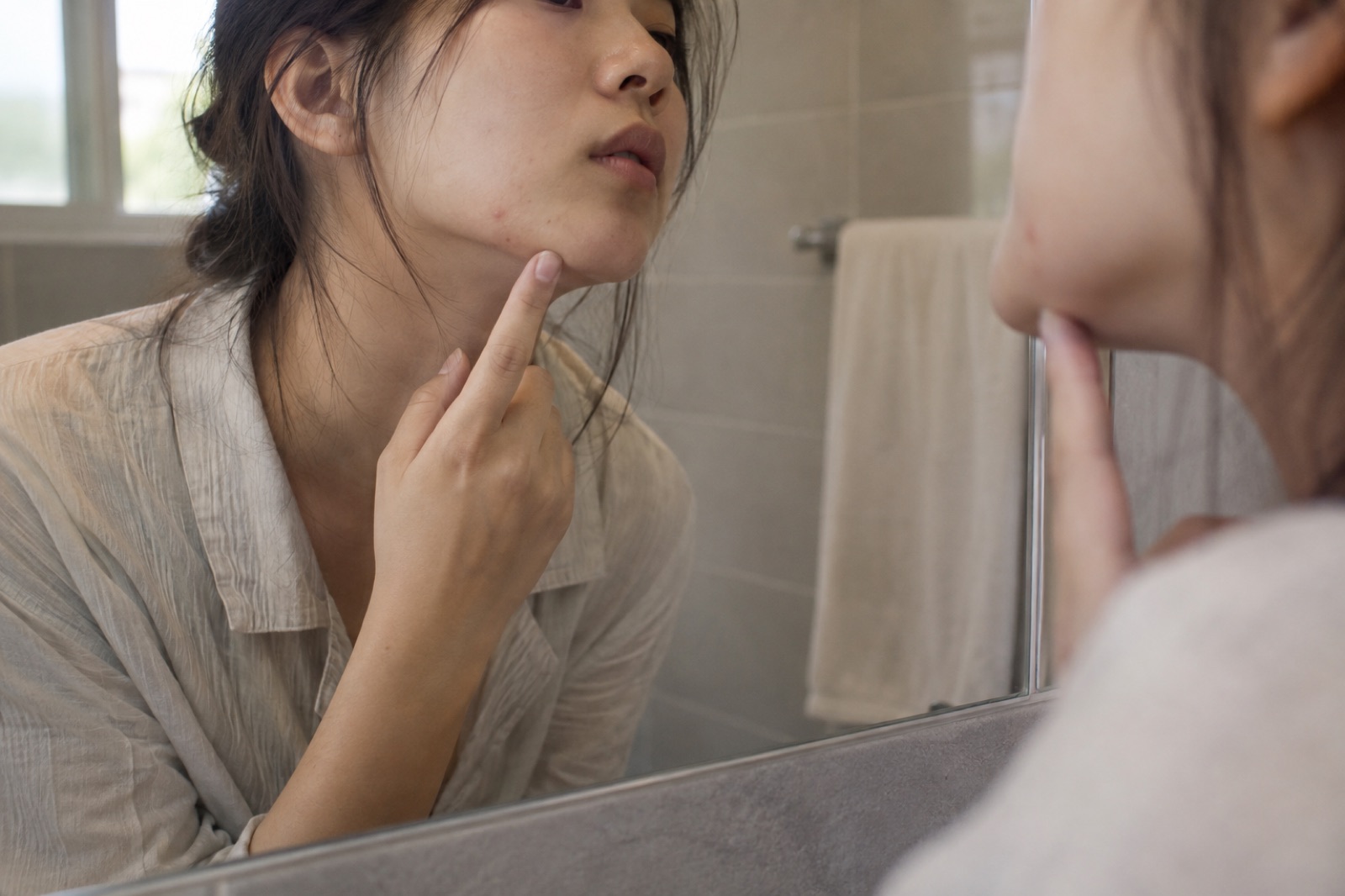 욕실 거울에 비친 여성이 검지로 턱 끝을 짚은 채 볼의 작은 붉은 점 하나를 들여다보는 모습