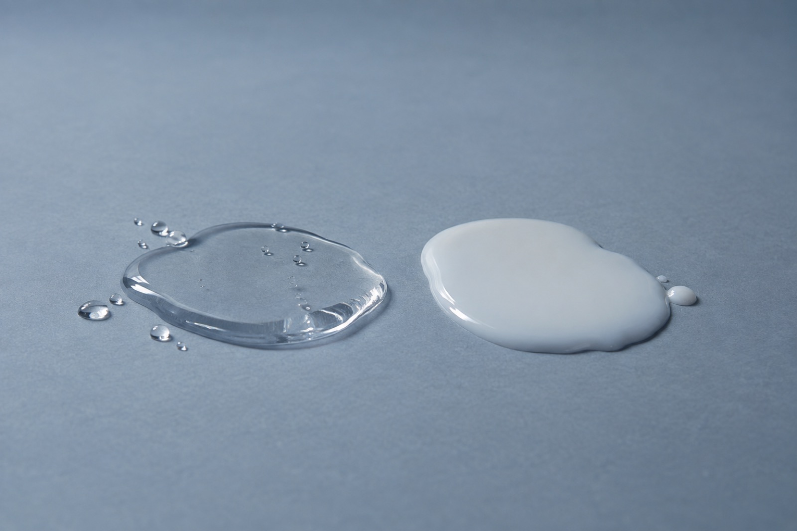 On a blue-gray background, clear liquid with splashed droplets on the left and a smooth pool of white milky lotion on the right