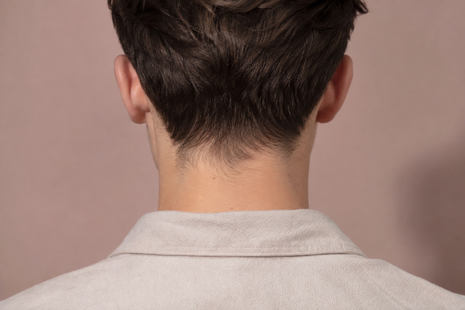 Likod na anyo sa harap ng pink na pader: maayos na gupit na hairline at makinis na batok sa ibabaw ng kuwelyo ng beige na polo