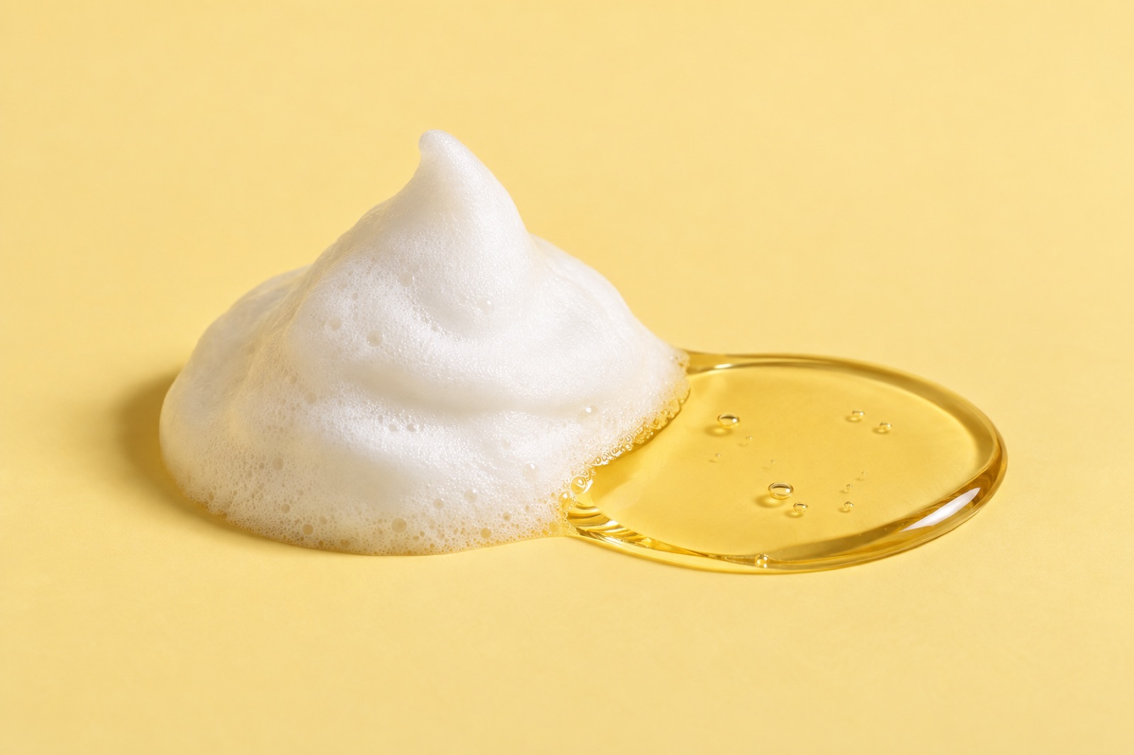 A soft mound of white foam on a yellow background next to a round, spreading pool of clear golden oil