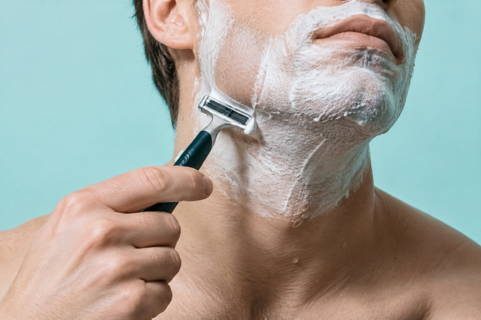 水色の背景の前で、あごと首に白い泡をつけた男性があごのラインの下をカミソリで剃っている様子