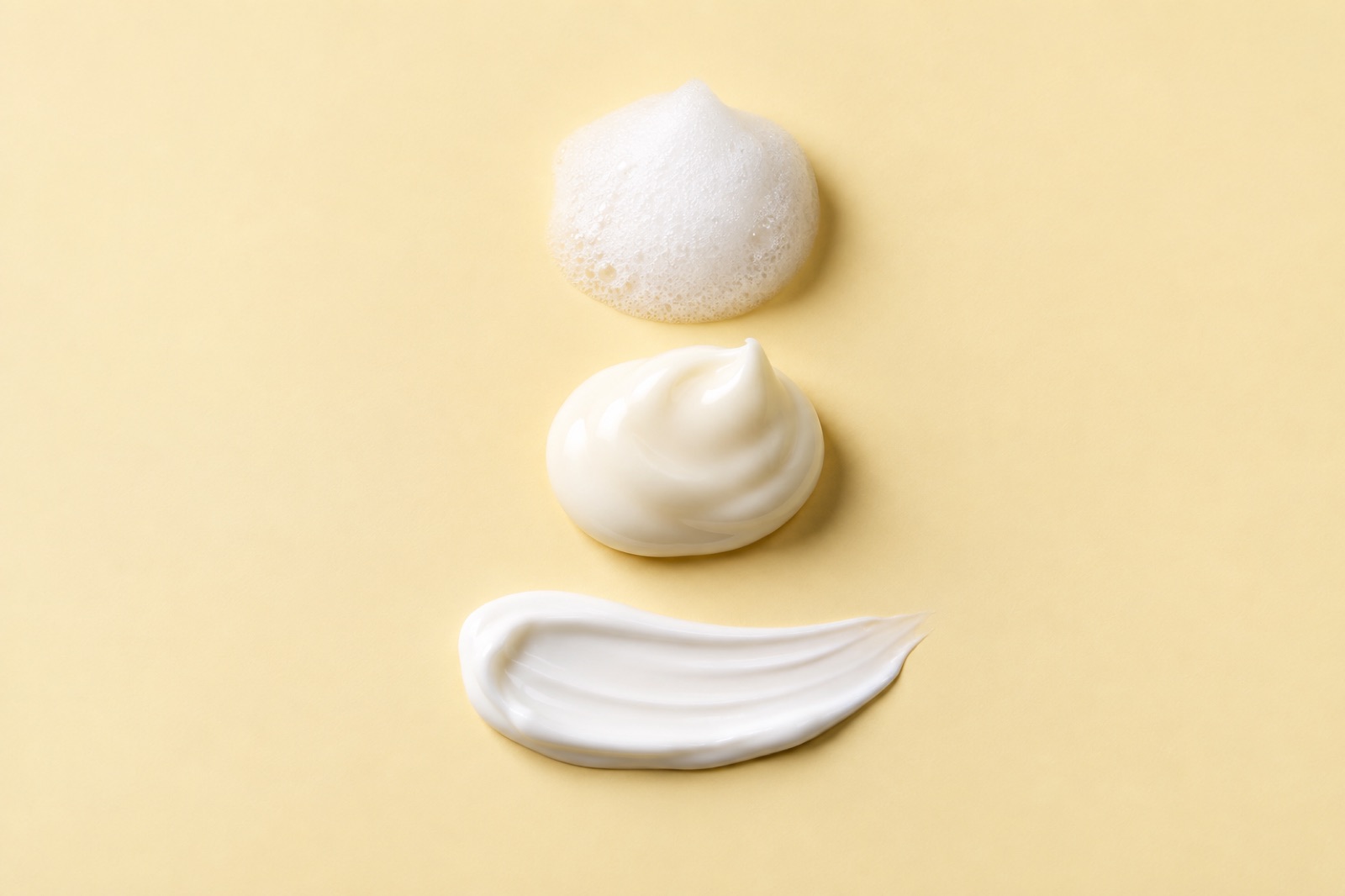 On a pale yellow background, from top to bottom: white foam, a rounded dollop of cream, and a long smear of white cream