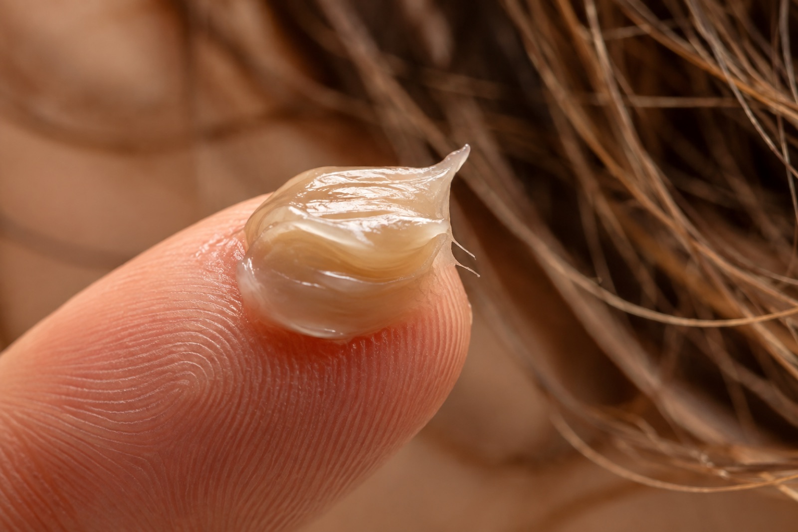 Malapitang kuha ng malinaw na parang cream na hair product sa dulo ng hintuturo, sa harap ng magulong buhok