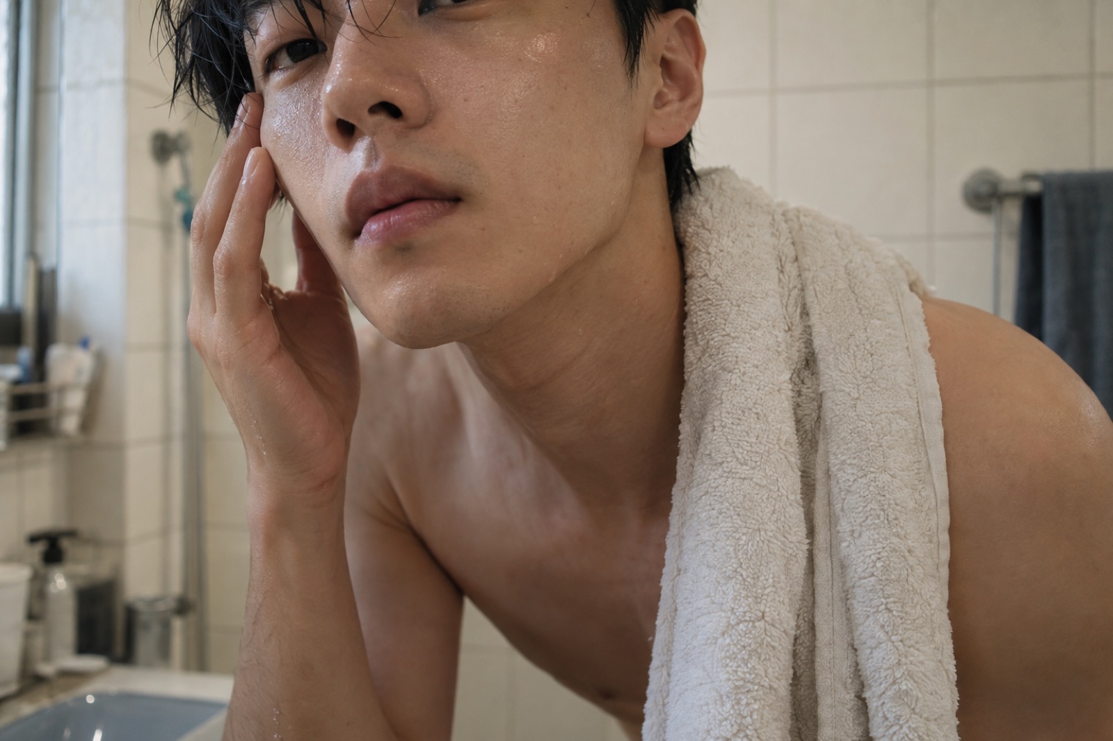 A man at a bathroom sink touching his wet face with a white towel draped over one shoulder