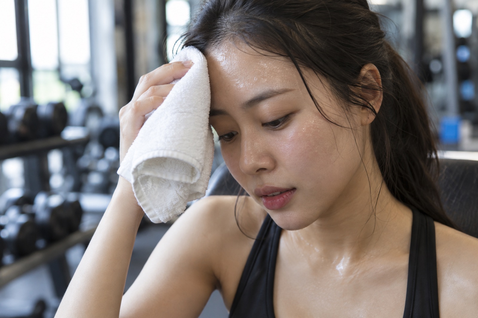 운동 기구가 흐릿하게 보이는 실내에서 땀이 맺힌 여성이 흰 수건을 이마에 대고 고개를 숙인 모습
